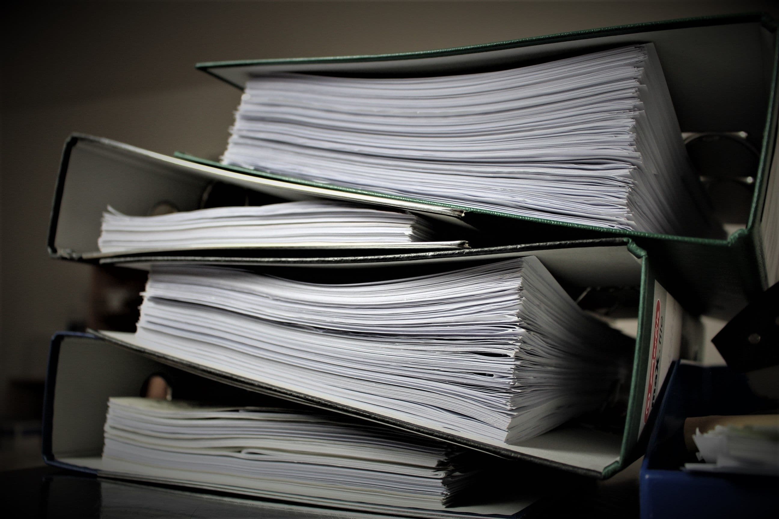 A stack of ring binders overflowing with paper documents.